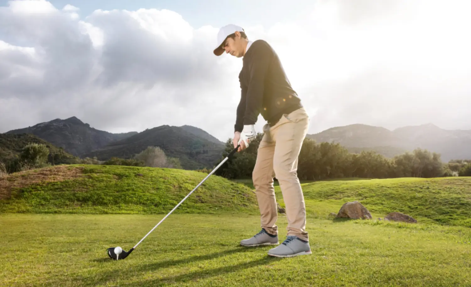 A golfer prepares to tee off on a lush green golf course, with mountains and clouds in the background under a bright sky